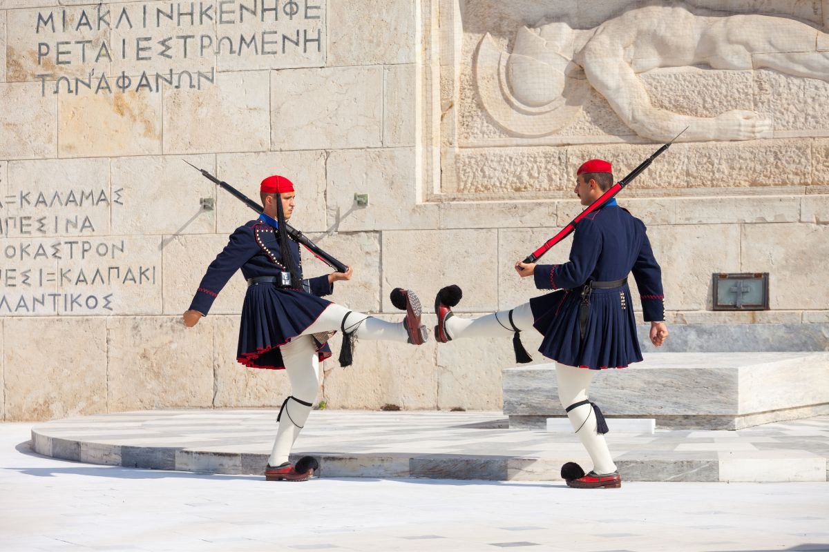 The Evzones, Greek presidential guards, perform the ceremonial changing of the guard in Athens, dressed in traditional uniform with red berets and carrying rifles, in front of a historic monument.